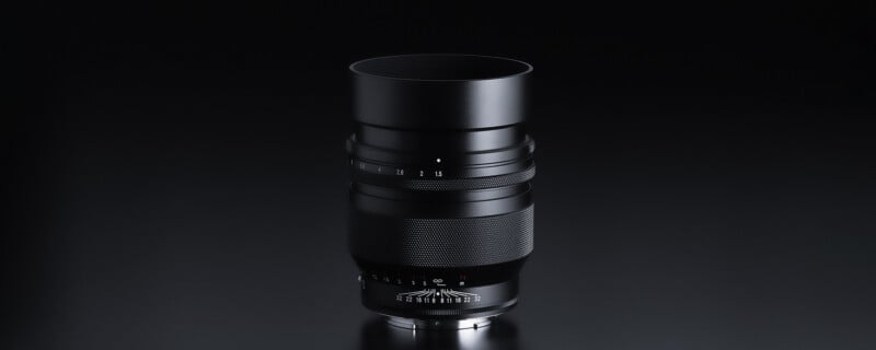 A black camera lens stands upright against a dark, reflective background. The barrel features various markings and adjustment rings, with a textured grip section in the middle. The lens hood is extended, enhancing the overall sleek and professional appearance.