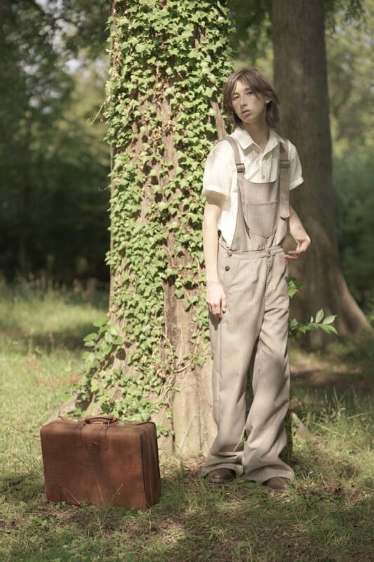 A person with long hair wearing light-colored overalls and a short-sleeve shirt leans against a tree covered in ivy in a forest. A vintage brown suitcase is placed on the ground beside them. The scene is calm and bathed in natural light.