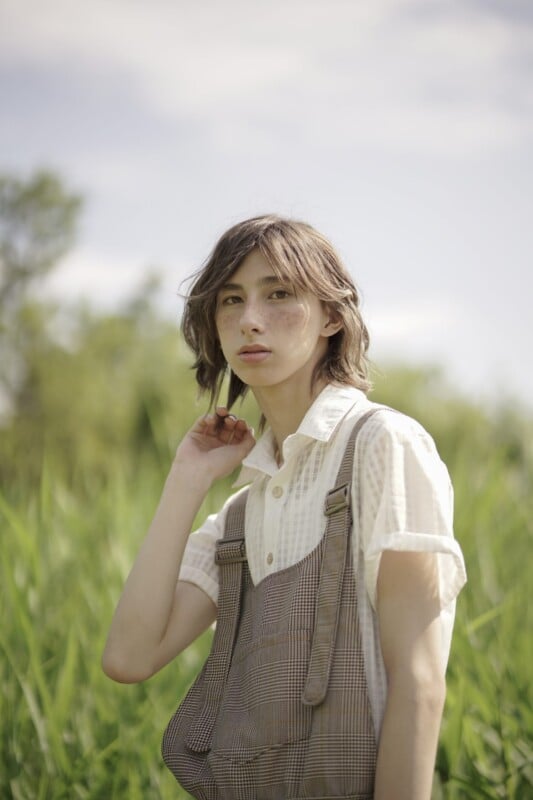 A person with medium-length hair stands outdoors in a grassy field. They are wearing a white collared shirt under brown plaid overalls and are looking directly at the camera. The background is slightly blurred, with greenery and a light blue sky visible.