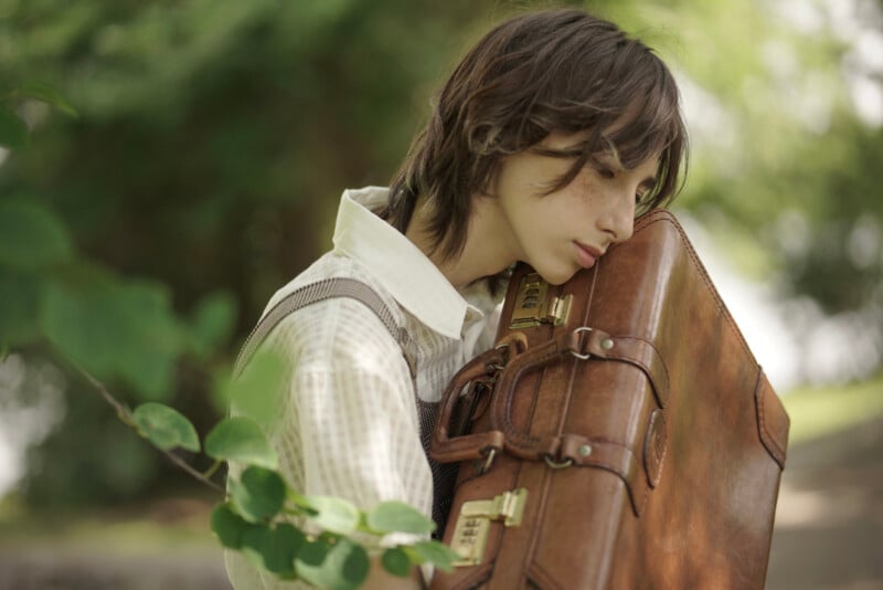 A person with chin-length brown hair, wearing a white shirt and vest, rests their head on a large, worn leather suitcase. They stand outdoors, surrounded by blurred greenery, conveying a sense of contemplation or fatigue.