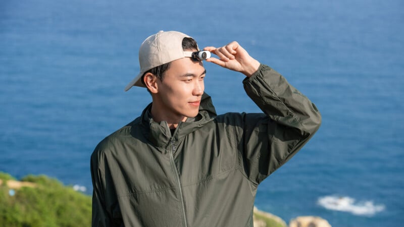 A person wearing a green jacket and a beige cap holds a small camera to their forehead while standing outdoors. The background features a body of water and a partially blurry shoreline with greenery.