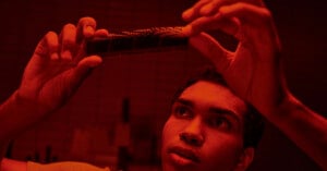 A person is examining a strip of photographic film held up to the light in a dim, red-lit room, likely a darkroom. Their expression appears focused and thoughtful as they inspect the film. The setting suggests a process related to photo development.