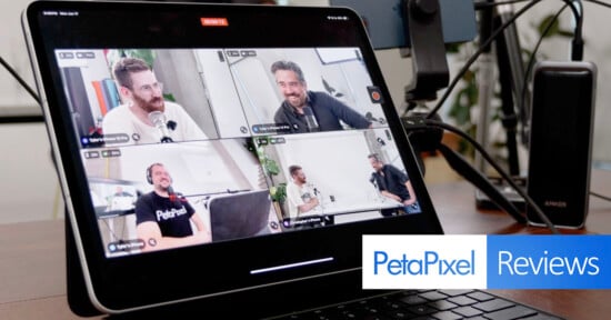 A tablet screen displays a video call with four men engaged in a discussion, each in their own quadrant. Visible behind the tablet are various electronic devices and cables. A "PetaPixel Reviews" logo is visible in the bottom right corner of the image.