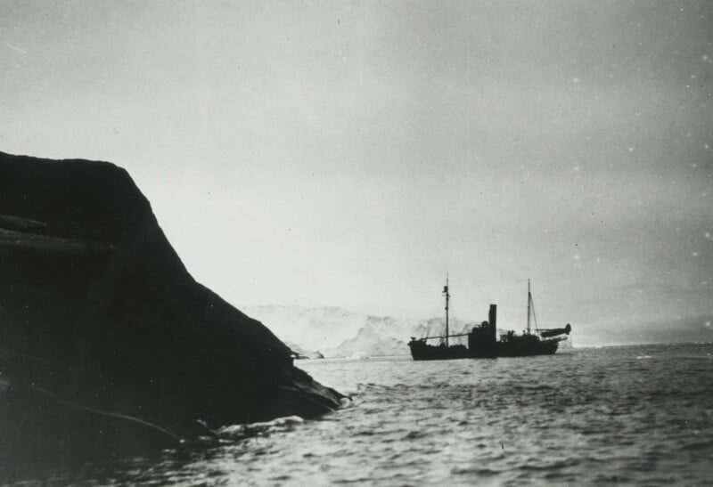 A black and white photo of a steam-powered ship in the distance, floating on calm waters near an iceberg or snow-covered landmass. In the foreground on the left, a cliff or large rock formation partially obscures the view. The sky appears overcast.