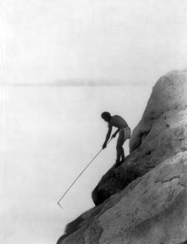 A person stands on a steep rocky ledge, leaning forward and holding a long, thin pole towards the water below. The background is mostly empty, giving a sense of height and isolation. The image is in black and white.