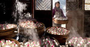 A woman stands in a dimly lit room, holding a tray with steaming round dishes. Several bamboo steamers filled with steaming rice topped with pink decorations are on large pots in the foreground. Wooden lattice screens and doors are in the background.