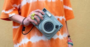 A person wearing an orange and white tie-dye shirt holds a gray Fujifilm Instax Mini 90 instant camera. They have a green scrunchie on their finger and several bracelets on their wrist, standing against a yellow background.
