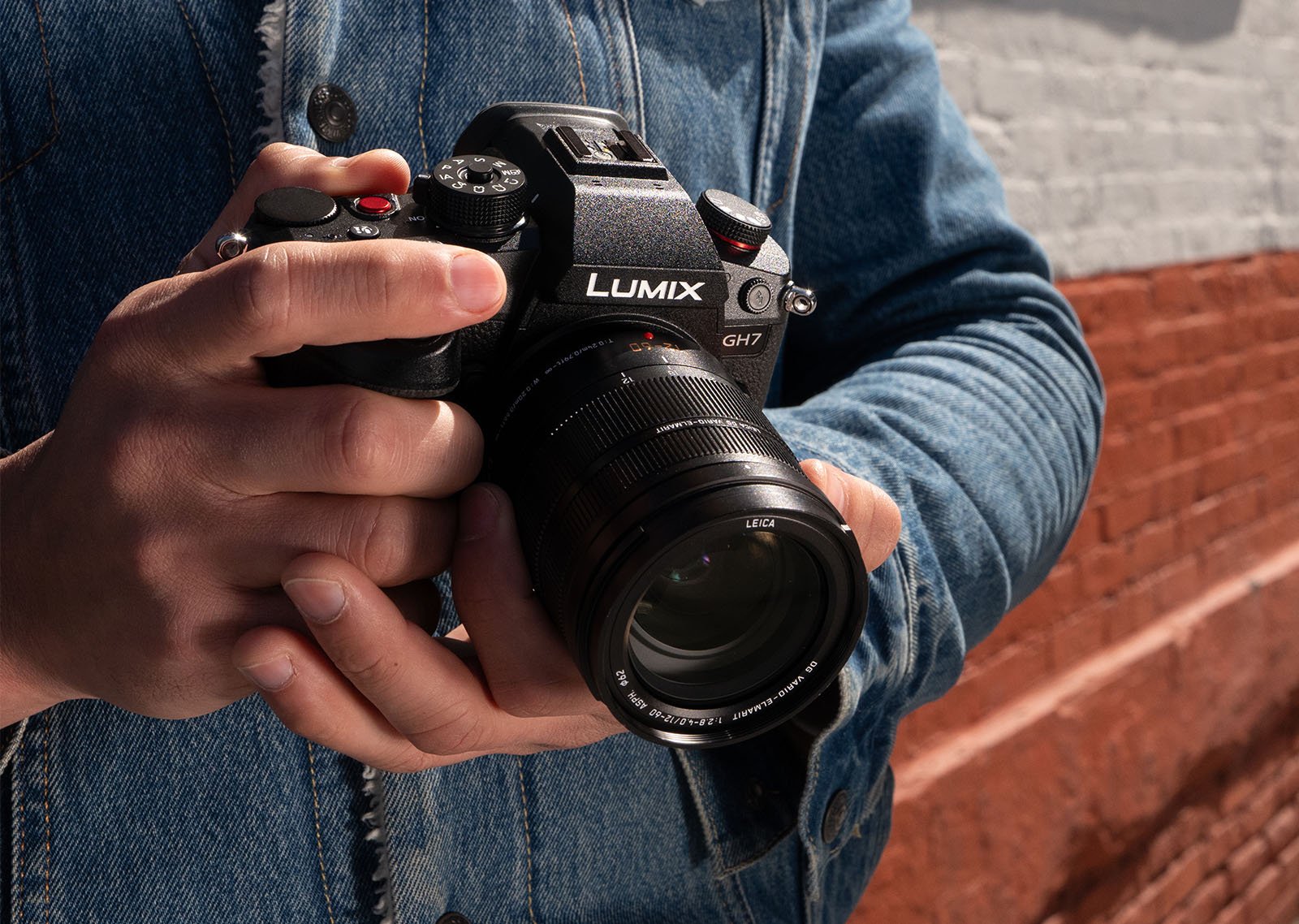 A person in a denim jacket holds a black Lumix GH7 camera with both hands against a background of a red and white brick wall. The camera is equipped with a large lens.