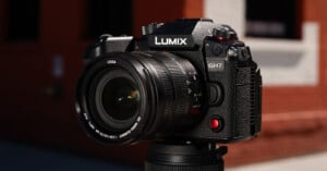 A Panasonic Lumix GH7 camera with a Leica lens mounted on a tripod, set against a slightly blurred brick building background. The camera's branding and controls are clearly visible.