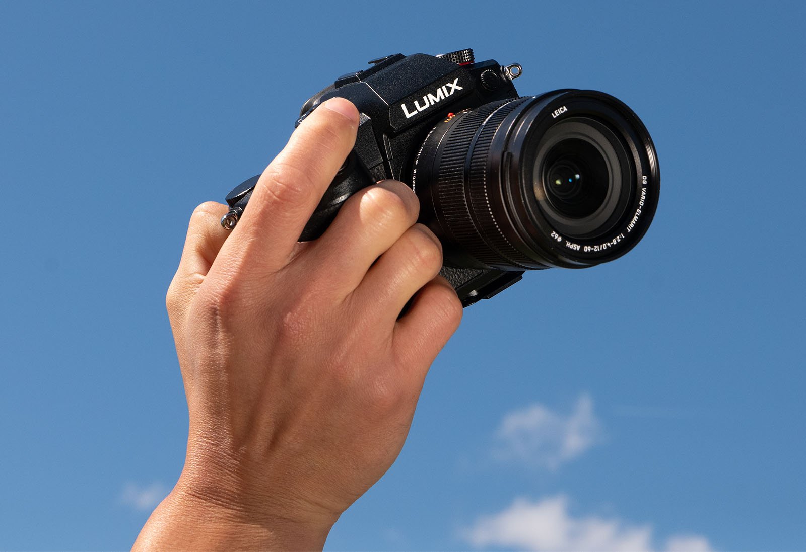 A hand holding a Panasonic Lumix camera against a clear blue sky with a few scattered clouds. The camera is pointed towards the viewer, showcasing its lens and front details.