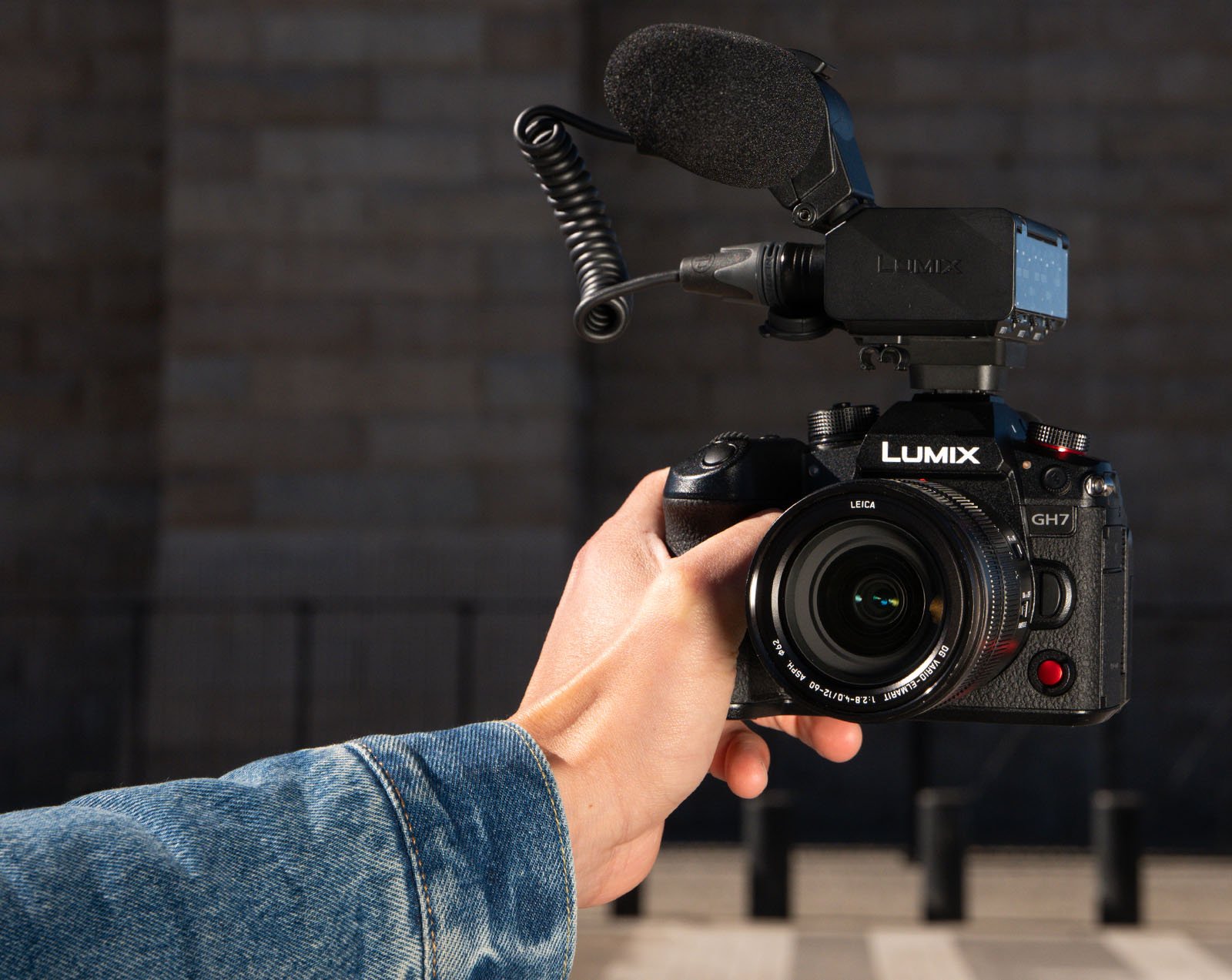 A person in a denim jacket holds up a Lumix GH7 camera with an attached microphone. The camera features prominent branding, visible lens elements, and control dials. The background is out of focus, showing a building and several black posts.