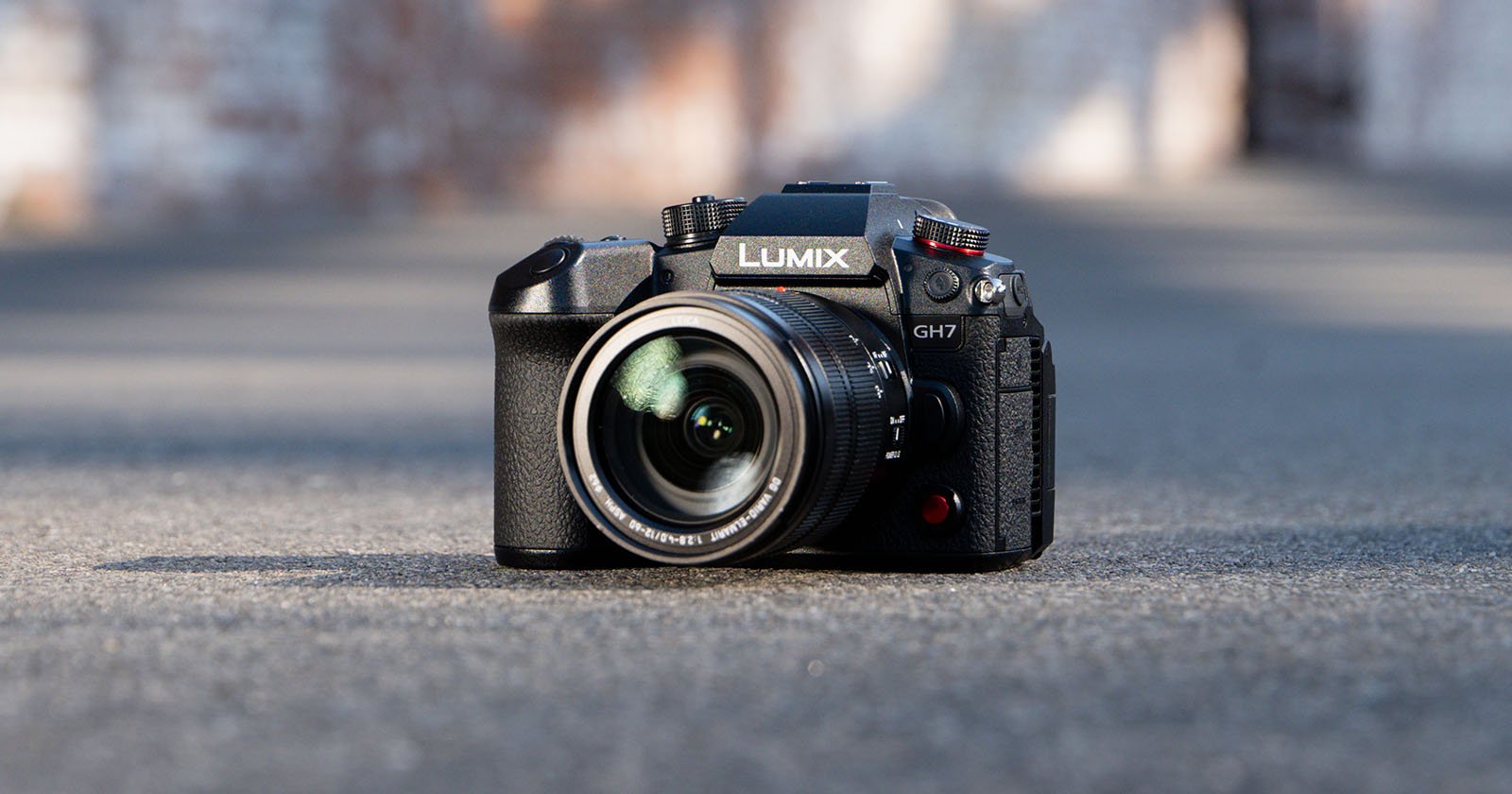 A Lumix GH7 camera is placed on a smooth gray surface. The camera lens is prominently visible, pointing towards the viewer, with the Lumix logo on the top. The background is blurred, highlighting the focus on the camera.