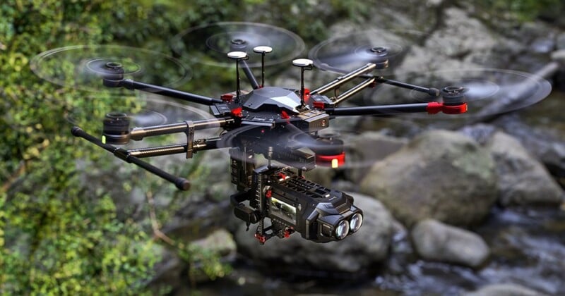 A black and red drone with multiple propellers is seen flying outdoors over rocky terrain and green foliage. The drone is equipped with a high-resolution camera and stabilization gear, suitable for aerial photography or videography.