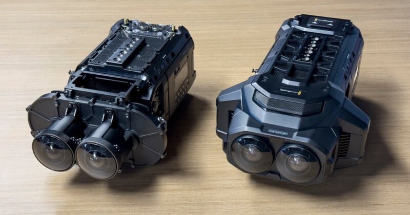 Two advanced dual-lens 3D cameras are placed on a wooden surface. The cameras are sleek and futuristic, with various buttons and dials on their bodies. One camera has an open compartment revealing internal components, while the other is fully assembled.