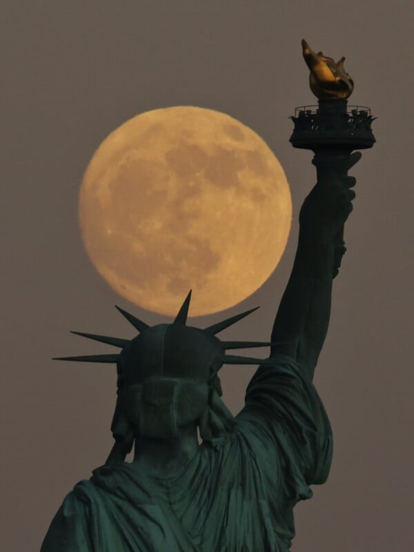 A full moon rises behind the Statue of Liberty, casting a dramatic glow around the monument's torch. The night sky is clear, allowing the golden moon to stand out prominently against the silhouette of the statue.