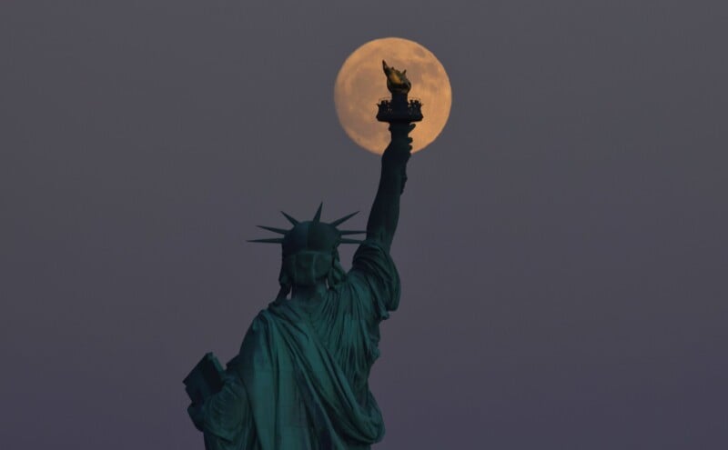 The Statue of Liberty is seen from behind with an illuminated full moon perfectly aligned with the torch she holds high in the sky. The scene is set against a dusk sky, creating a dramatic and captivating view.
