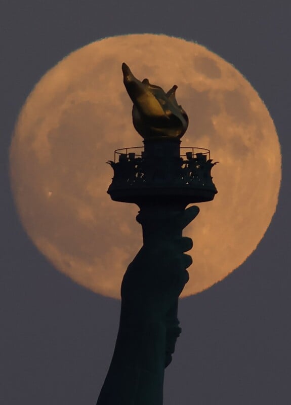 A large, bright, full moon sets behind the torch of the Statue of Liberty in this dramatic silhouette. The torch appears in the foreground with the intricate details visible, creating a striking contrast against the glowing backdrop of the moon.
