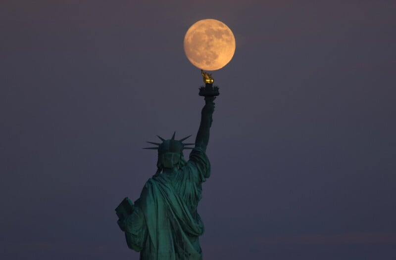 The Statue of Liberty is seen from behind, holding its torch aloft with a full moon perfectly positioned above it, creating the illusion that the moon is being held by the statue. The sky is dusky, adding a dramatic and serene atmosphere to the scene.