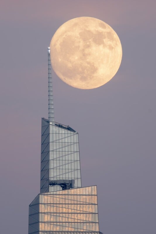 A large, glowing full moon is seen resting atop the spire of a modern glass skyscraper during dusk, creating an optical illusion. The building reflects the soft twilight, while the moon's luminous surface stands out against the dusky sky.