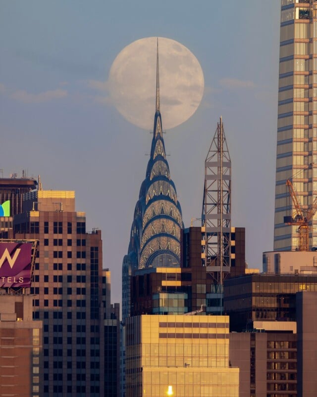 The image features the iconic Chrysler Building in New York City, with the full moon aligned perfectly behind its spire. The surrounding skyscrapers and construction cranes are illuminated by the evening light, creating a captivating cityscape.
