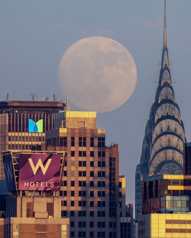A full moon rises over a cityscape featuring the iconic art deco Chrysler Building and a high-rise with a large purple "W Hotels" sign. The moon appears large in the twilight sky, creating a striking backdrop to the buildings.