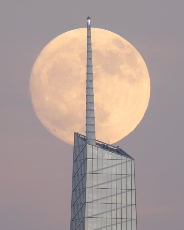 The image shows the full moon rising behind the spire of a modern glass skyscraper, creating an optical illusion where the spire appears to be puncturing the moon. The sky is a dusky twilight shade, giving the scene a serene, ethereal quality.