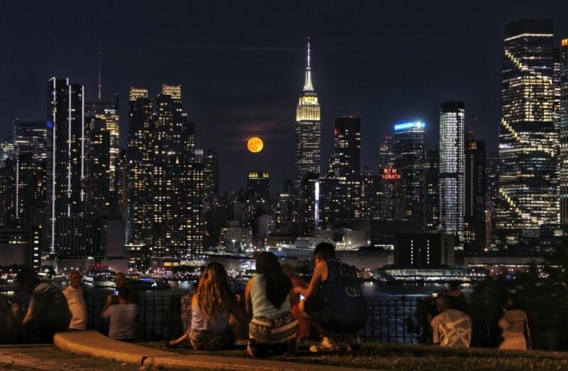 A group of people sit and look across the Hudson River at the illuminated New York City skyline at night. The Empire State Building, along with other skyscrapers, stands tall, while a full moon rises in the background, creating a picturesque and vibrant cityscape.