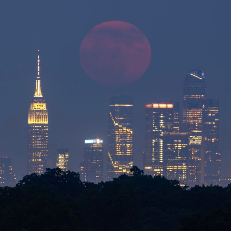 A large, full moon with a reddish hue rises over the New York City skyline at dusk. Prominent skyscrapers, including the Empire State Building and other illuminated high-rises, stand out against the darkening sky. Trees are visible at the bottom of the image.