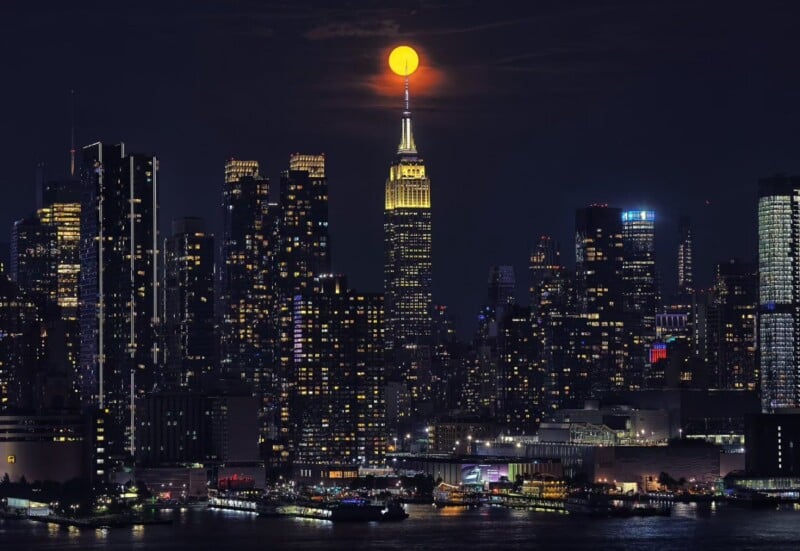 A nighttime cityscape of New York City, featuring a prominent skyline with the Empire State Building at the center. A large, bright moon appears to be perched atop the building's spire, illuminating the surrounding skyscrapers and reflecting over the water below.