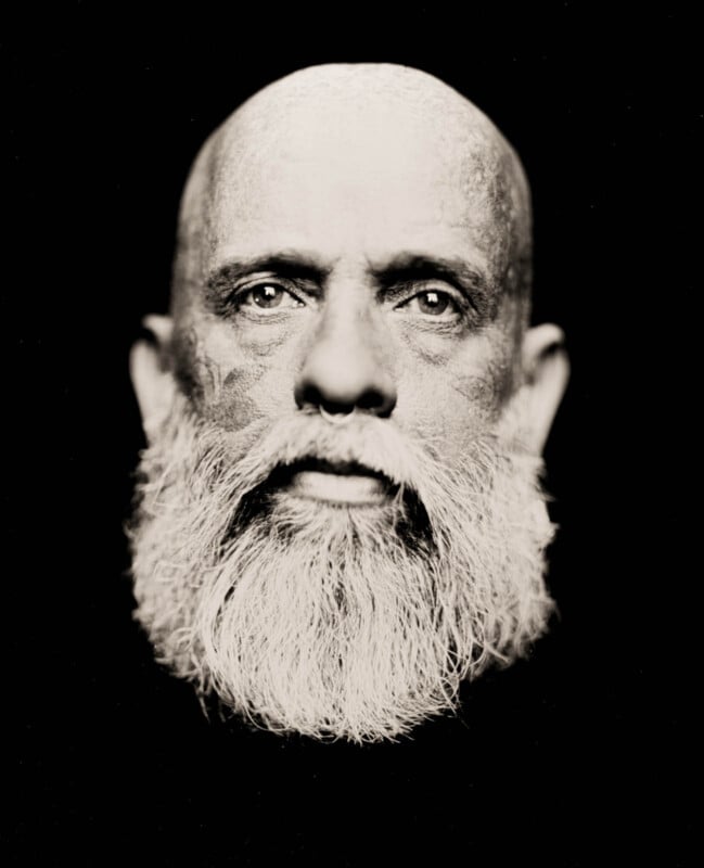 Black-and-white close-up portrait of a bald man with a full, white beard. The background is black, and the spotlight highlights his contemplative expression and facial details.