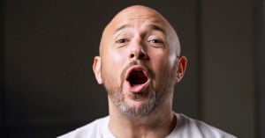 A bald man with a short beard is shown from the shoulders up against a neutral background. He has his mouth open wide as if he is speaking or singing. He is wearing a light-colored shirt.