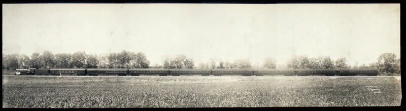 A panoramic black-and-white photograph of a long passenger train moving through a rural area. The train, consisting of multiple cars, travels on tracks bordered by open fields and trees in the background. The sky is clear and bright, indicating a sunny day.