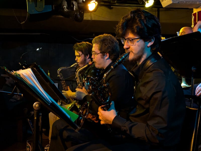 Three musicians are playing saxophones in a dimly lit area, possibly a concert or rehearsal space. They are seated in a row, each holding sheet music, and appear focused on their performance. The background includes music stands and stage lights overhead.