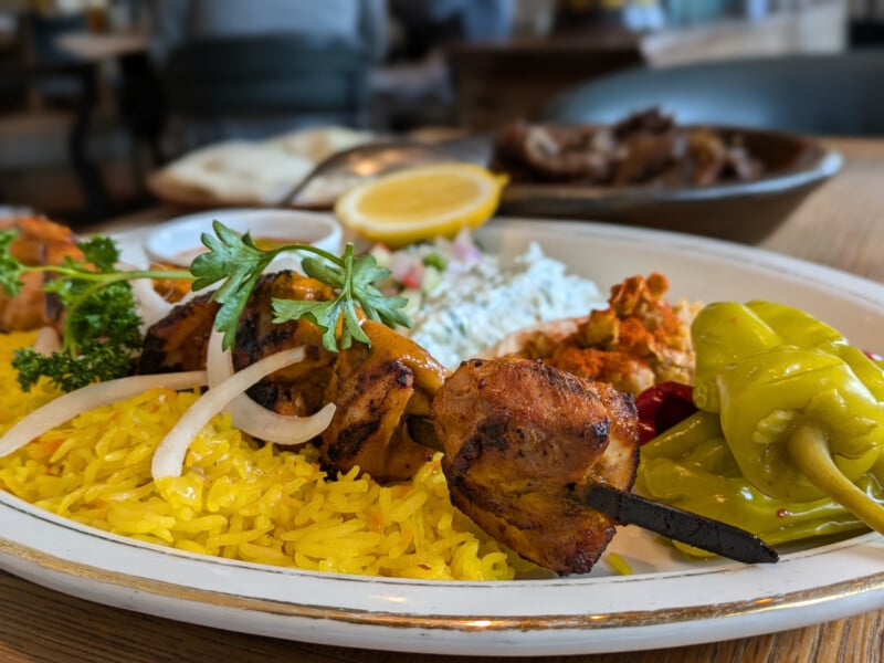 A plate of vibrant, yellow saffron rice is topped with grilled chicken skewers, garnished with fresh parsley and onions. The dish also includes roasted peppers, a lemon wedge, and sides such as a creamy sauce and vegetables, all beautifully presented on a white plate.