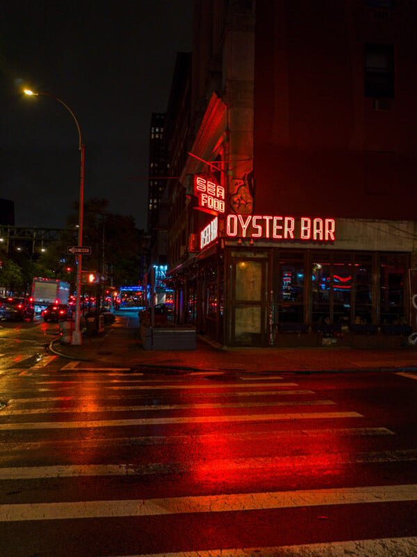 A dimly lit street at night with a neon-lit sign that reads "OYSTER BAR" and "SEA FOOD" in bright red. The reflections of the lights glimmer on the wet pavement, and there are cars and buildings visible in the background, under a cloudy sky.