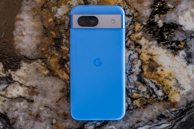 A blue smartphone with a black camera module is placed on a marbled surface. The phone features a central "G" logo, indicating it is a Google Pixel device. The marbled background has a mix of black, white, and brown colors.