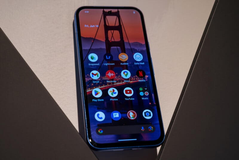 A smartphone with a home screen displaying various app icons, including Gmail, Google Maps, YouTube, Netflix, Google Photos, and others. The background image on the screen features the Golden Gate Bridge during a sunset. The date on the phone reads Fri, Jun 14.