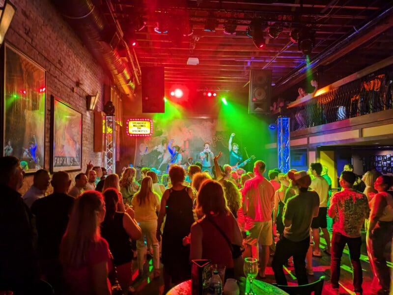 A crowded nightclub pulses with vibrant red and green lights as a live band performs on stage. People are dancing and enjoying the music, with some gathering near the bar. The venue has a second-floor balcony with more spectators.