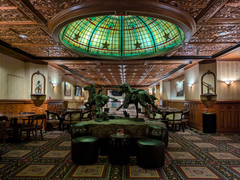 An opulent interior features an intricately decorated ceiling with a large stained glass dome. Below, a detailed sculpture of horses stands on a central display. Surrounding the sculpture are several tables and chairs set on a patterned carpet.