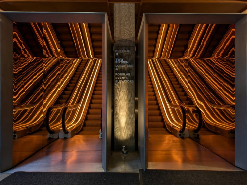 A modern staircase with wooden steps and warm, golden lighting that creates an optical illusion of multiple reflections. Two escalators are visible on either side of the stairs, framed by mirrors, enhancing the reflective effect. Central signboard instructions are visible.