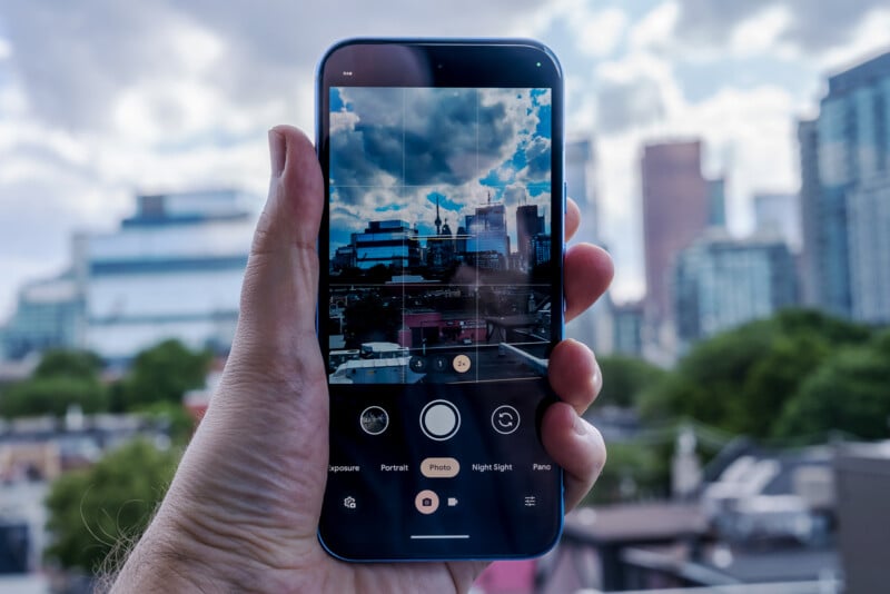 A hand holds a smartphone in camera mode, capturing a cityscape with tall buildings against a cloudy sky. The phone screen shows the same cityscape, framed and ready for a photograph. The background consists of a slightly blurred view of the city.