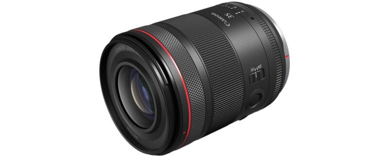 A black Canon RF 35mm f/1.8 IS Macro STM lens is shown against a white background. The lens features a wide focus ring, a control ring, and an image stabilization switch. It has a red ring near the front element, indicating its L-series status.