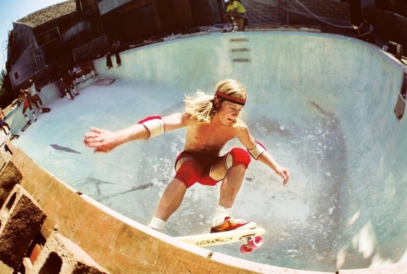 A skateboarder with long blonde hair, wearing a headband, red shorts, knee pads, and elbow pads, performs a trick on the edge of an empty swimming pool. Onlookers watch from the background. The pool's surface is worn and weathered.