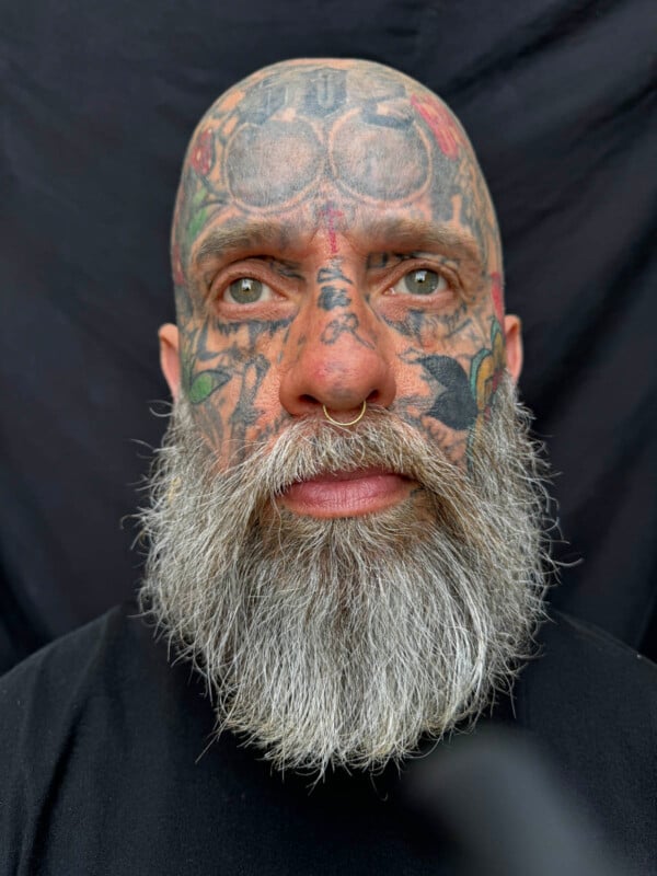 A man with a full gray beard, numerous facial tattoos, and a nose ring is shown against a dark background. He has a serious expression, and his tattoos cover his scalp, forehead, cheeks, and upper neck. He is wearing a plain black shirt.