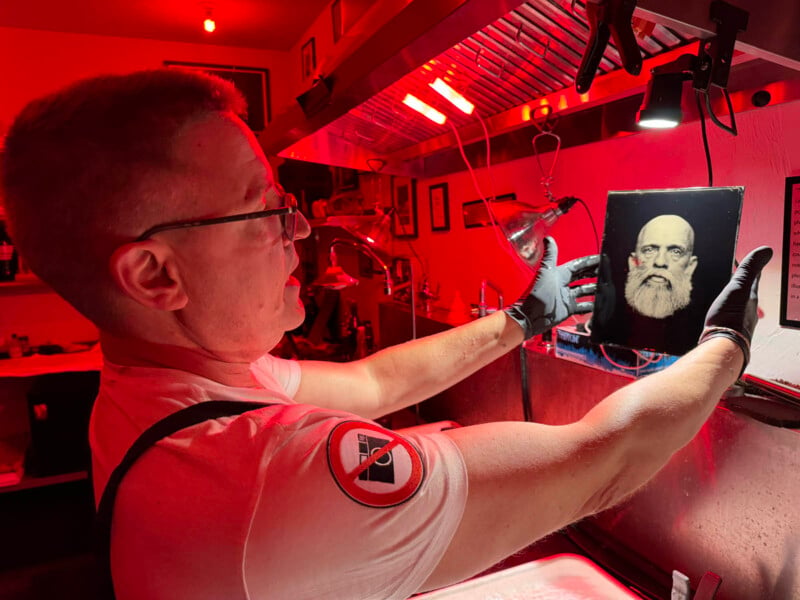 A person wearing gloves and a white shirt with a red and black patch is holding up and examining a photograph of an older bearded man under a red light in a dark room. The room appears to be a photography darkroom with various equipment and tools.