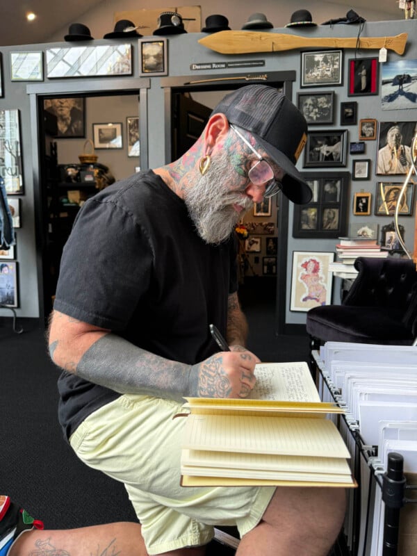 A bearded man with tattoos, wearing a black cap, glasses, a black t-shirt, and beige shorts, leans over a journal, writing intently. He is seated in a room filled with framed pictures and memorabilia on the walls.