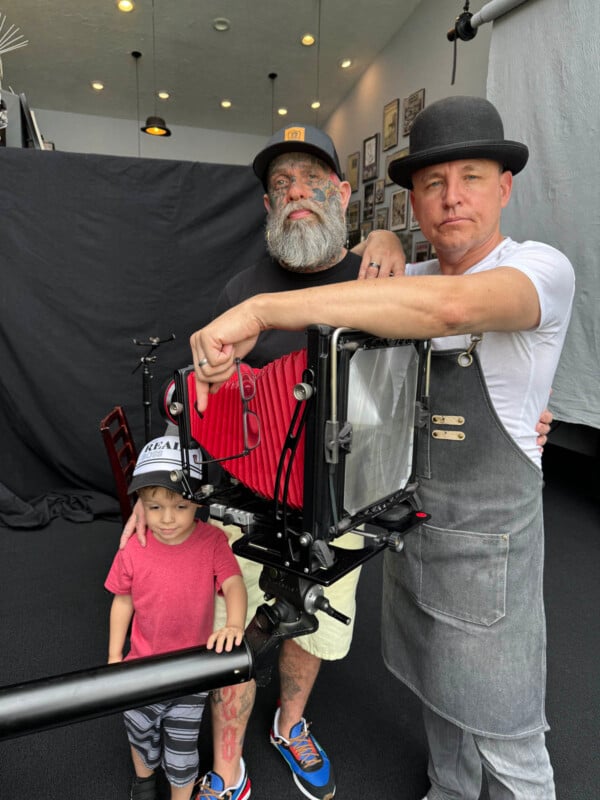 Two men and a young boy pose with a large vintage camera. The man on the left has tattoos and a beard. The man on the right wears a bowler hat and a gray apron. The young boy is wearing a red T-shirt and a baseball cap, standing in front of the man on the left.