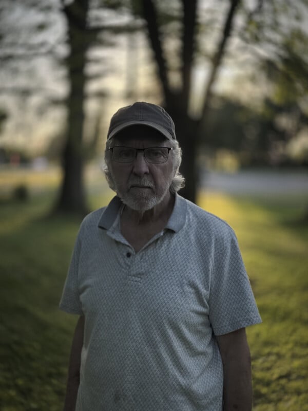 An elderly man with a gray beard, wearing glasses, a cap, and a light-colored polo shirt stands outside. The background is a blurry scene of trees and a road, with soft sunlight filtering through the leaves. The atmosphere feels calm and serene.