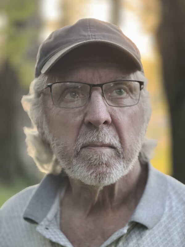 A close-up portrait of an elderly man with white hair and a beard wearing glasses and a brown baseball cap. He is looking slightly upwards with a thoughtful expression. The background is blurred, showing outdoor greenery and warm light.