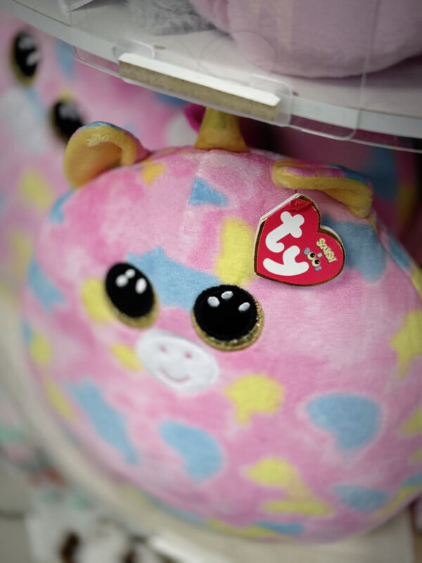 A close-up of a colorful, plush giraffe toy with large, expressive eyes. The giraffe is predominantly pastel pink with patches of blue, yellow, and green. It has yellow plush horns, ears, and a red Ty tag hanging from its ear.